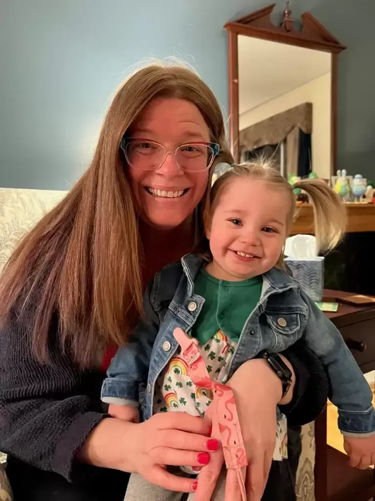 Woman with glasses holds smiling toddler; both looking at camera. Indoor setting, light skin tones.