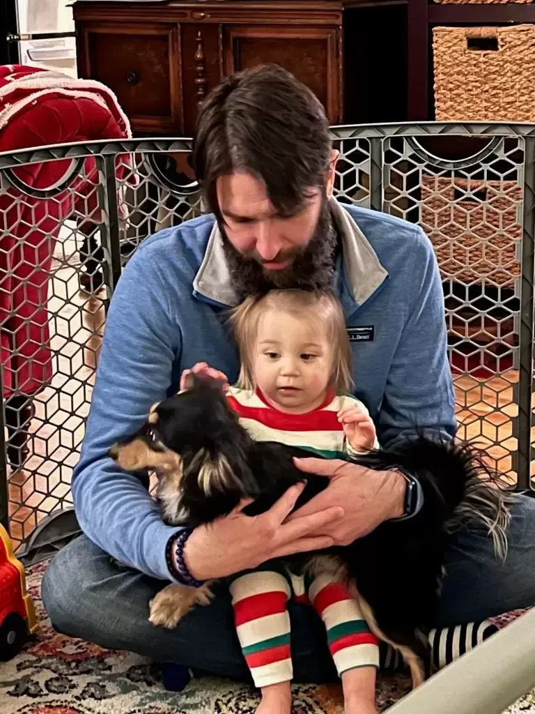 Man holding baby and dog, sitting inside a baby gate. Baby wears striped pajamas.