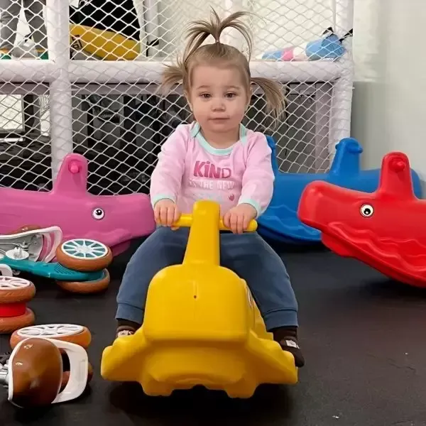 A young child with pigtails sits on a yellow ride-on toy in a playroom.