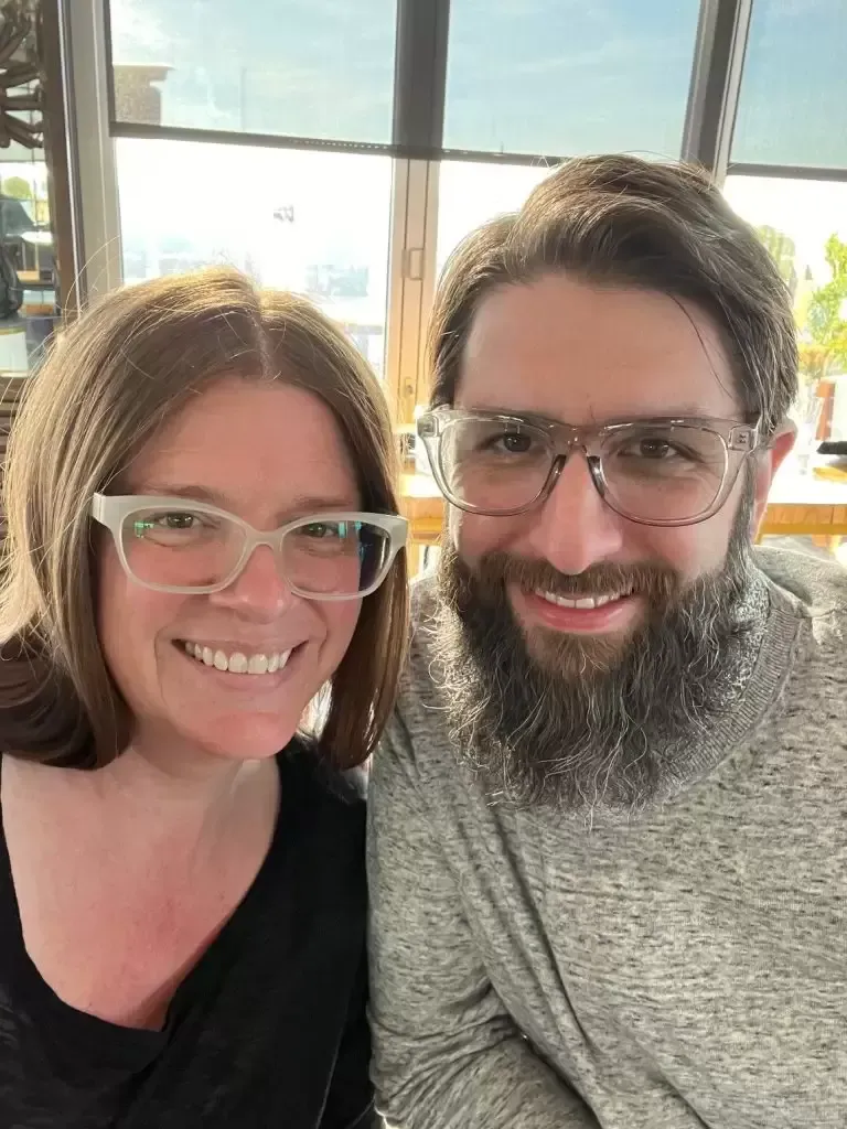 Woman and man wearing glasses smile, sitting inside near windows.