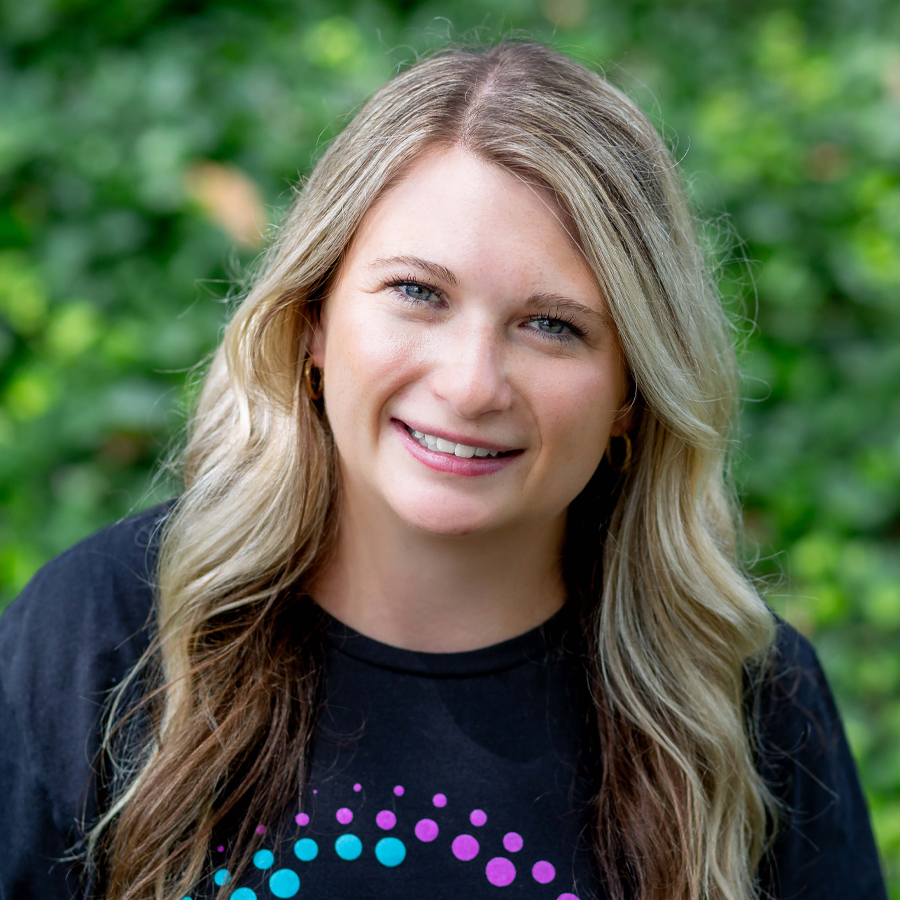 Woman with blonde hair smiles at the camera, wearing a black shirt with colorful design, outdoors.