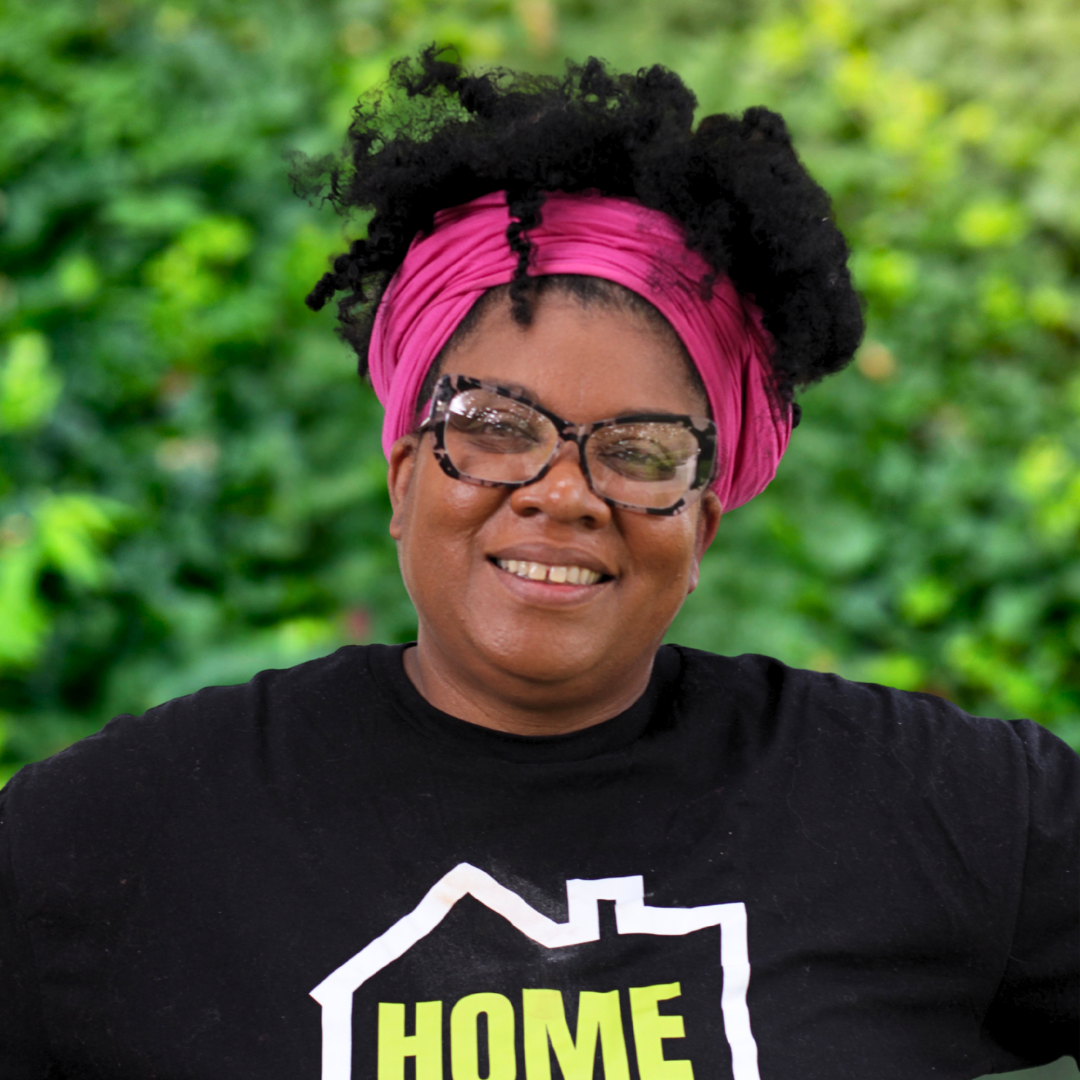 Smiling person with glasses and pink headwrap wearing a black HOME shirt outdoors