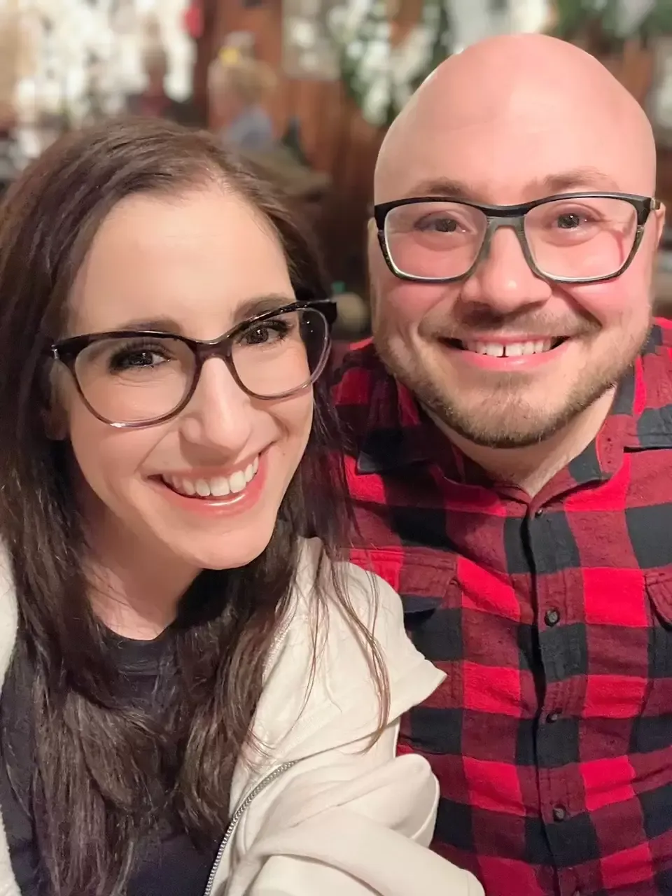 Smiling couple wearing glasses; woman with long hair, man with a bald head, in a cozy indoor setting.