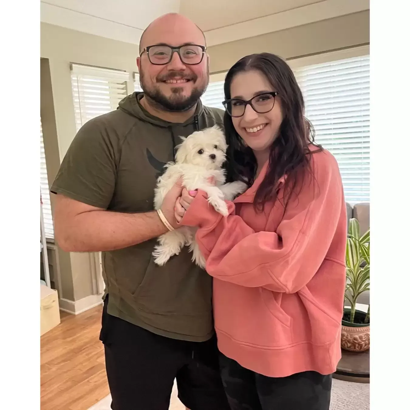 Man and woman holding a small, white puppy. They are smiling in a room with blinds and a plant.