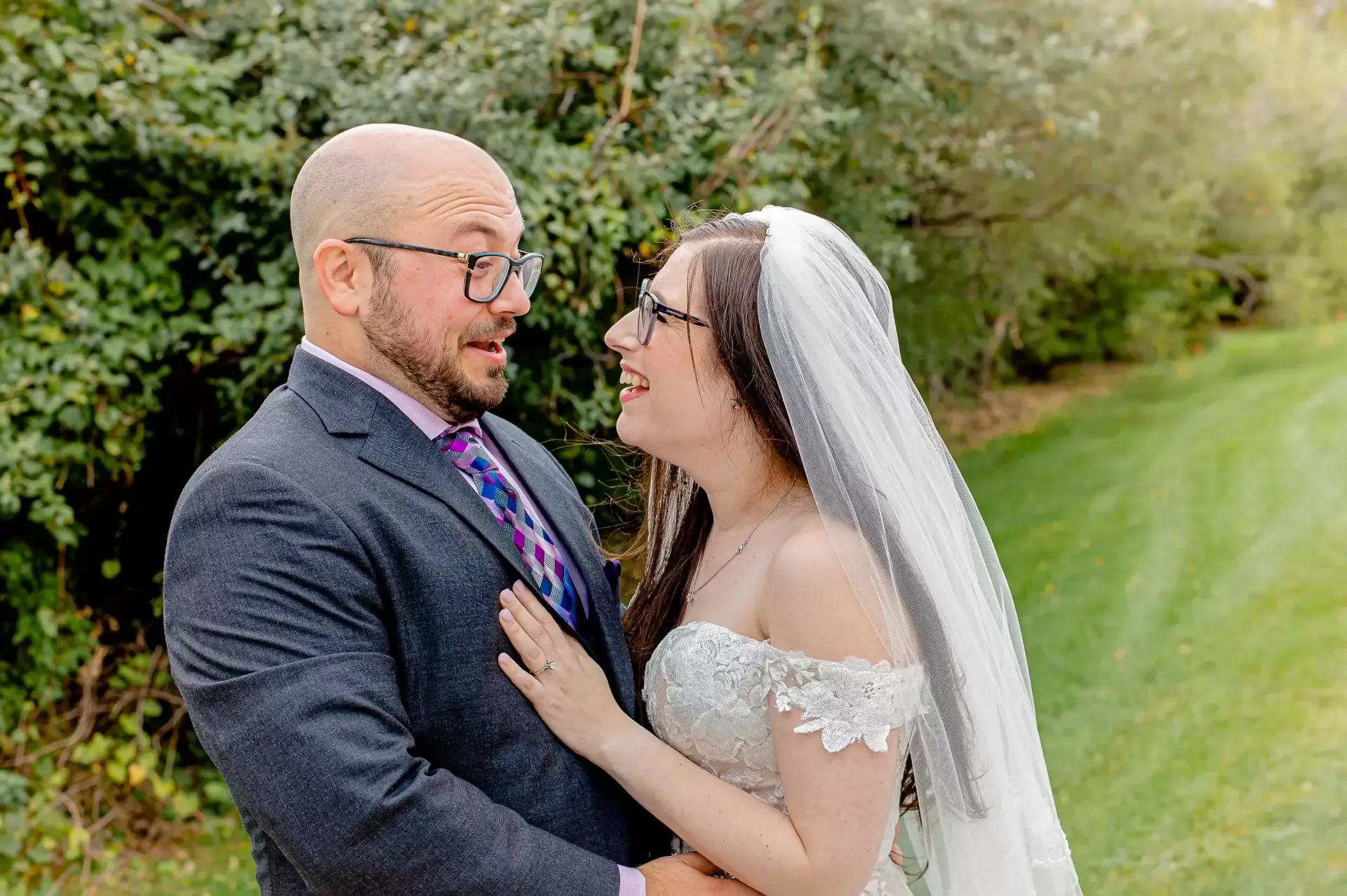 Bride and groom gaze at each other lovingly in outdoor setting. Bride wears white dress and veil. Groom in suit and glasses.
