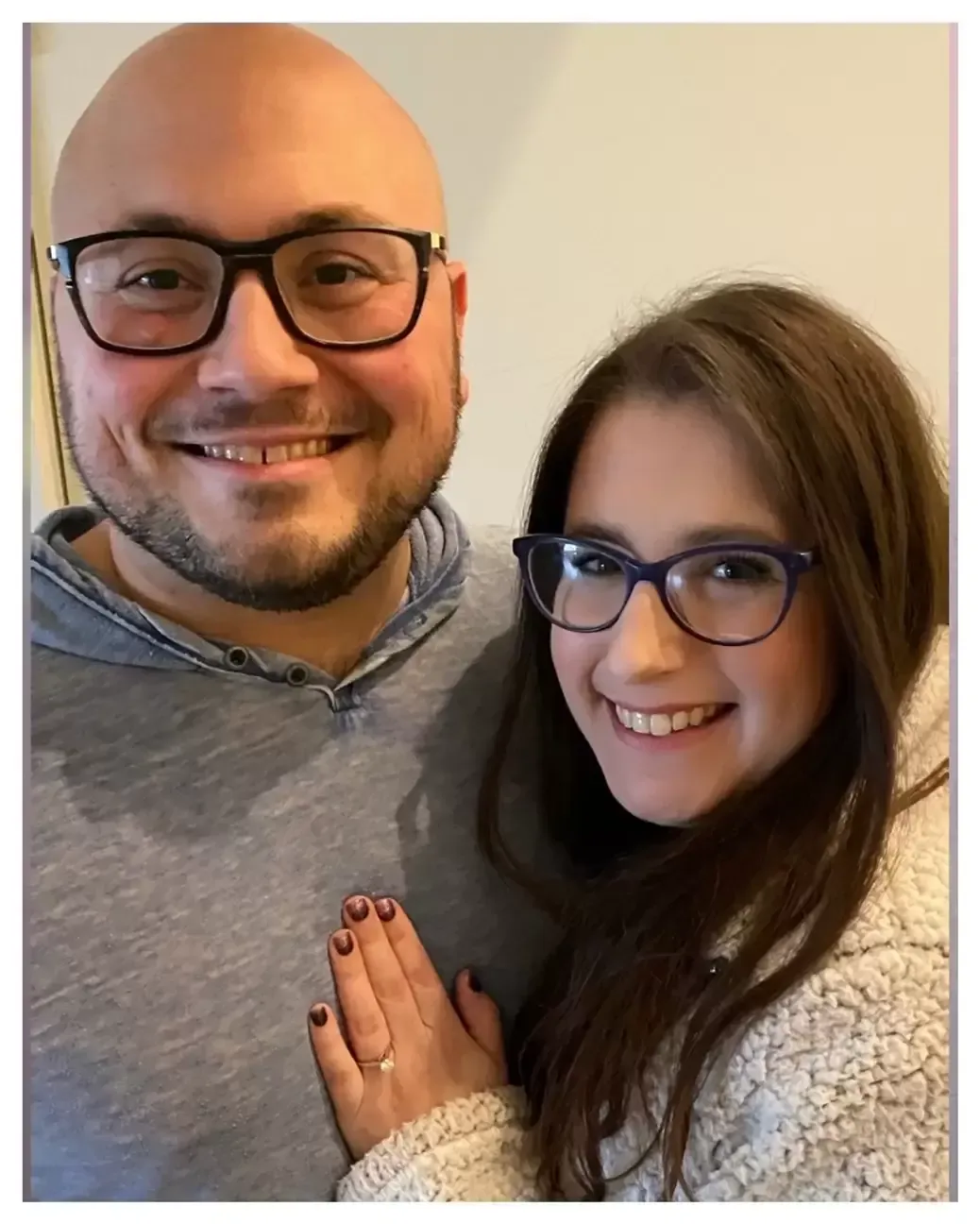 Man and young woman with glasses smiling, arms together, in a home setting. Man wearing gray hoodie, woman in white coat.