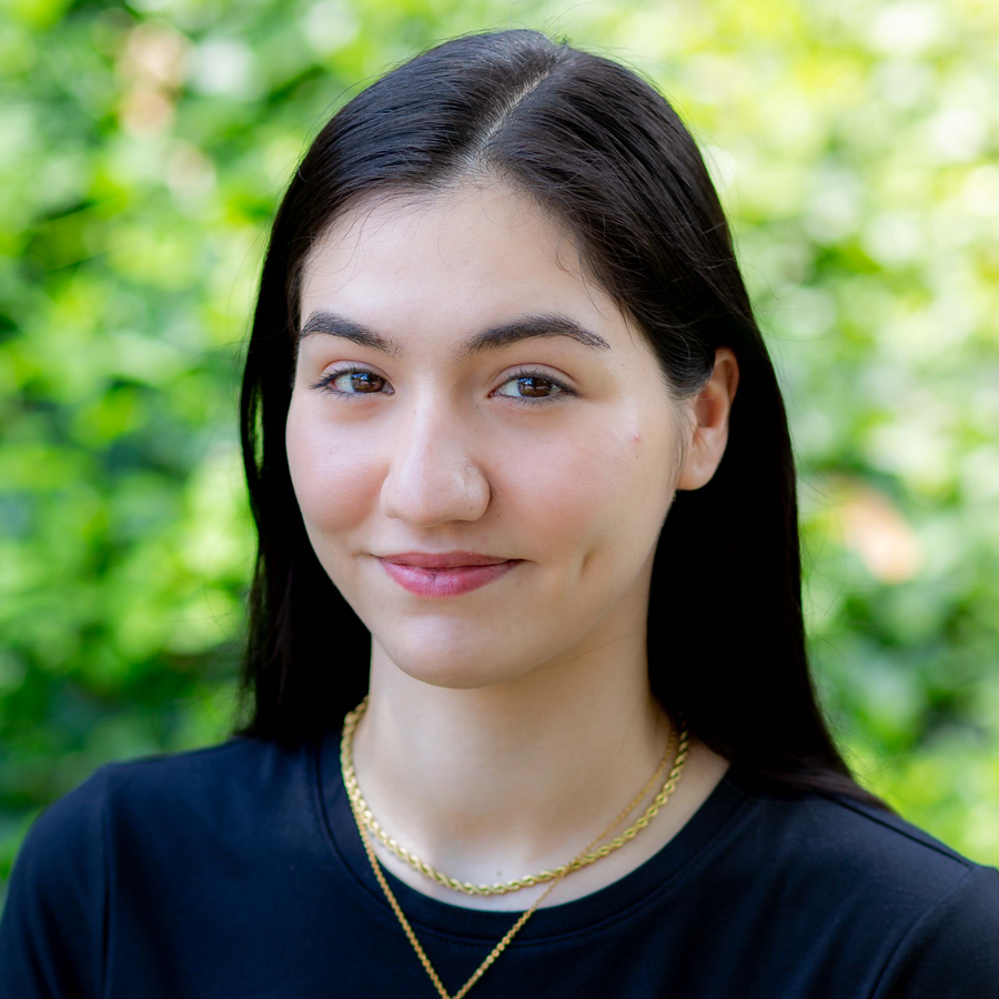 Woman with long brown hair and bangs smiles at the camera, wearing a black shirt in front of a white wall.