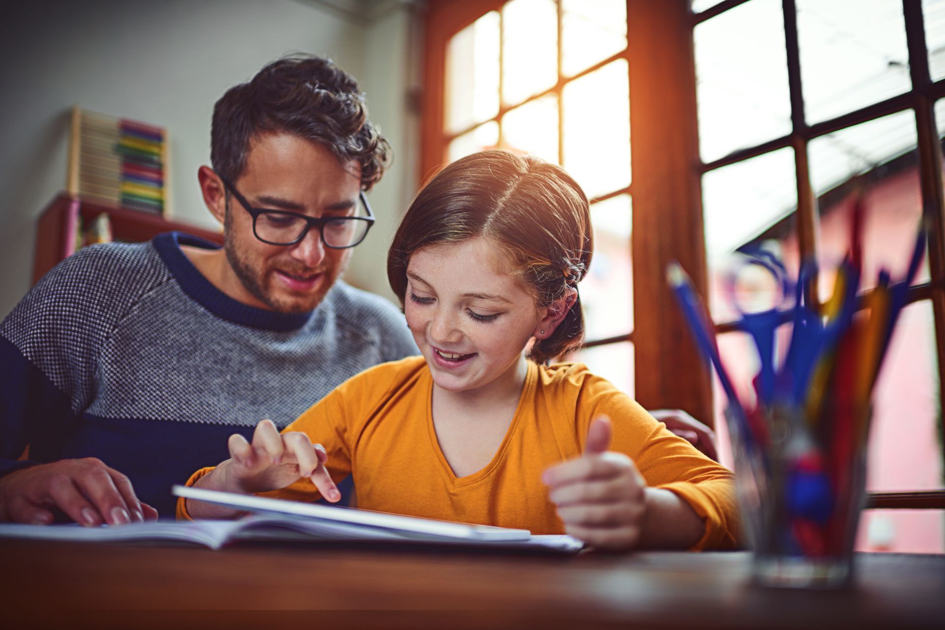 Man with glasses helps a girl with schoolwork at a desk near a window, both looking at a tablet.