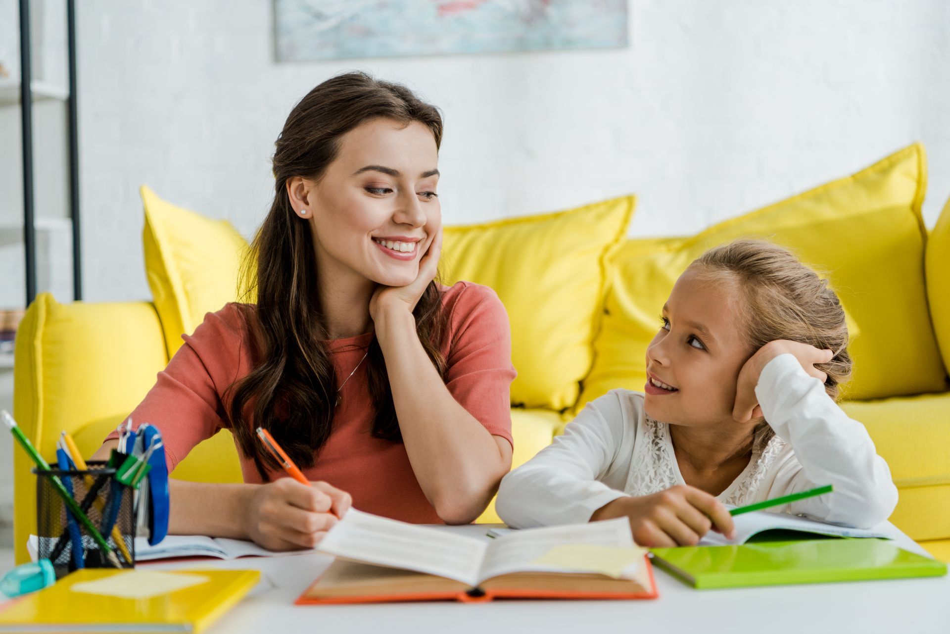 Woman smiles at girl as they write at a table, yellow couch background.