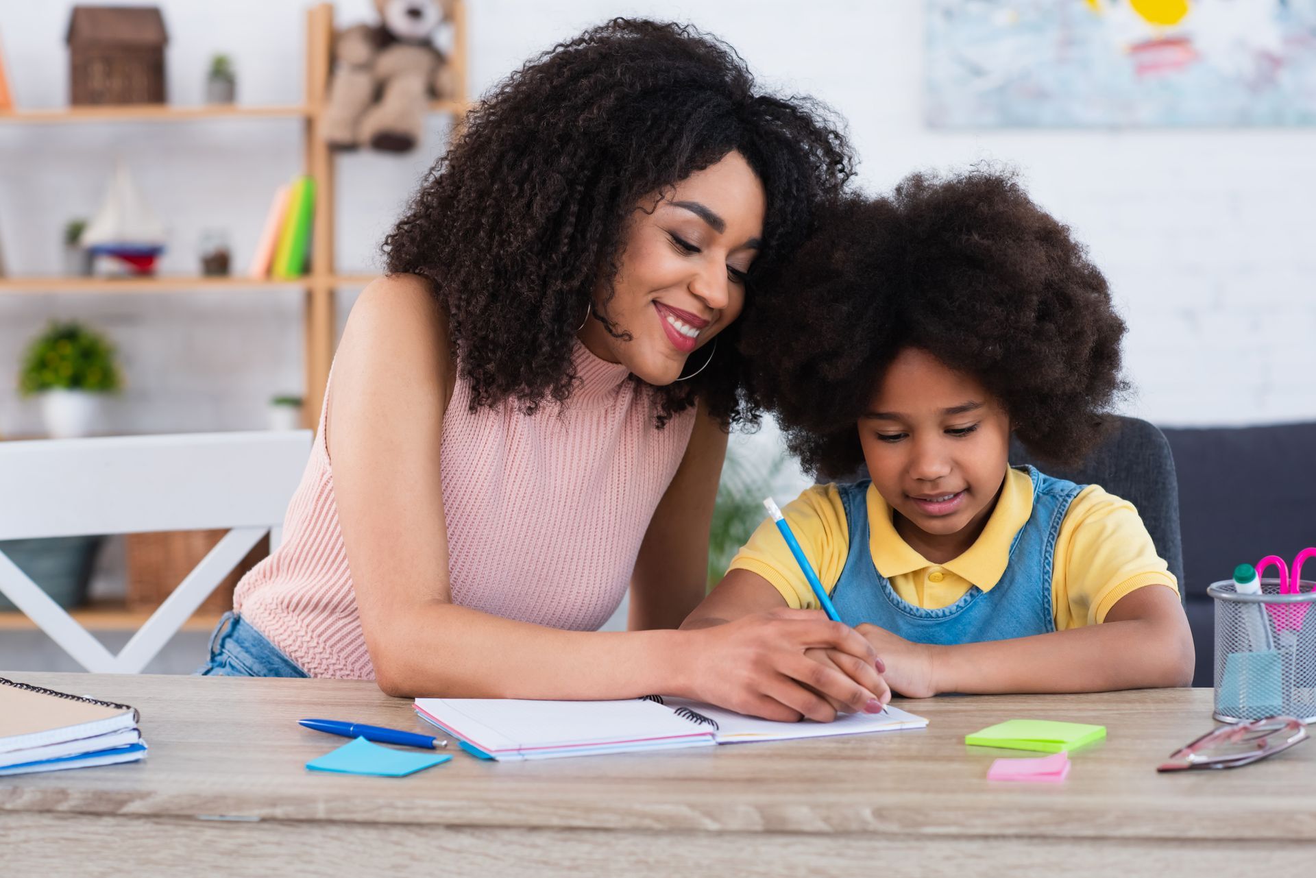 Woman smiles while helping child write at a desk. Notebook, pens, and phone visible.
