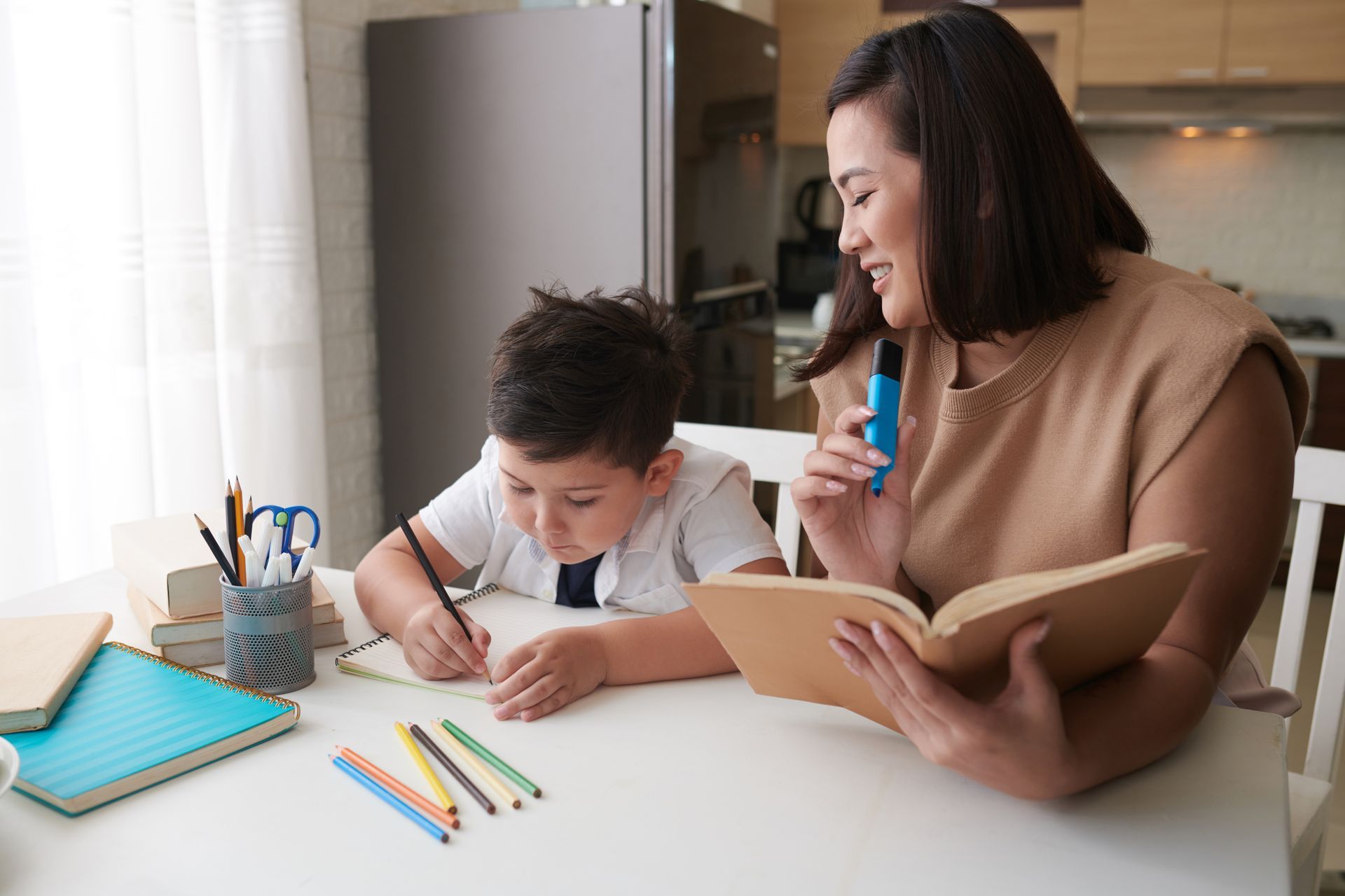 Woman helping a child write at a table.  She holds a book and a highlighter.  Notebooks and pencils are on the table.