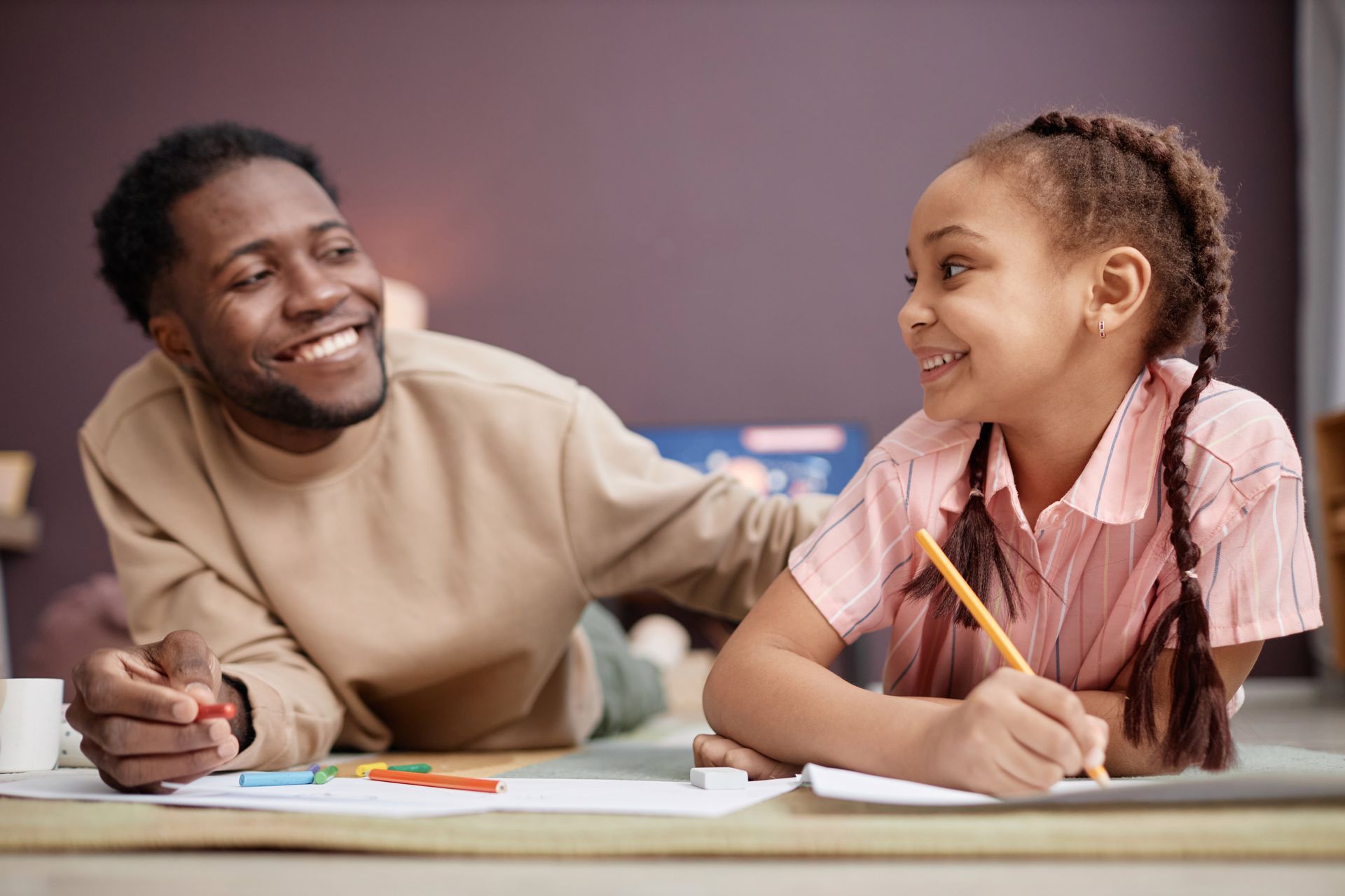 Man smiles at a girl as they draw together on the floor.