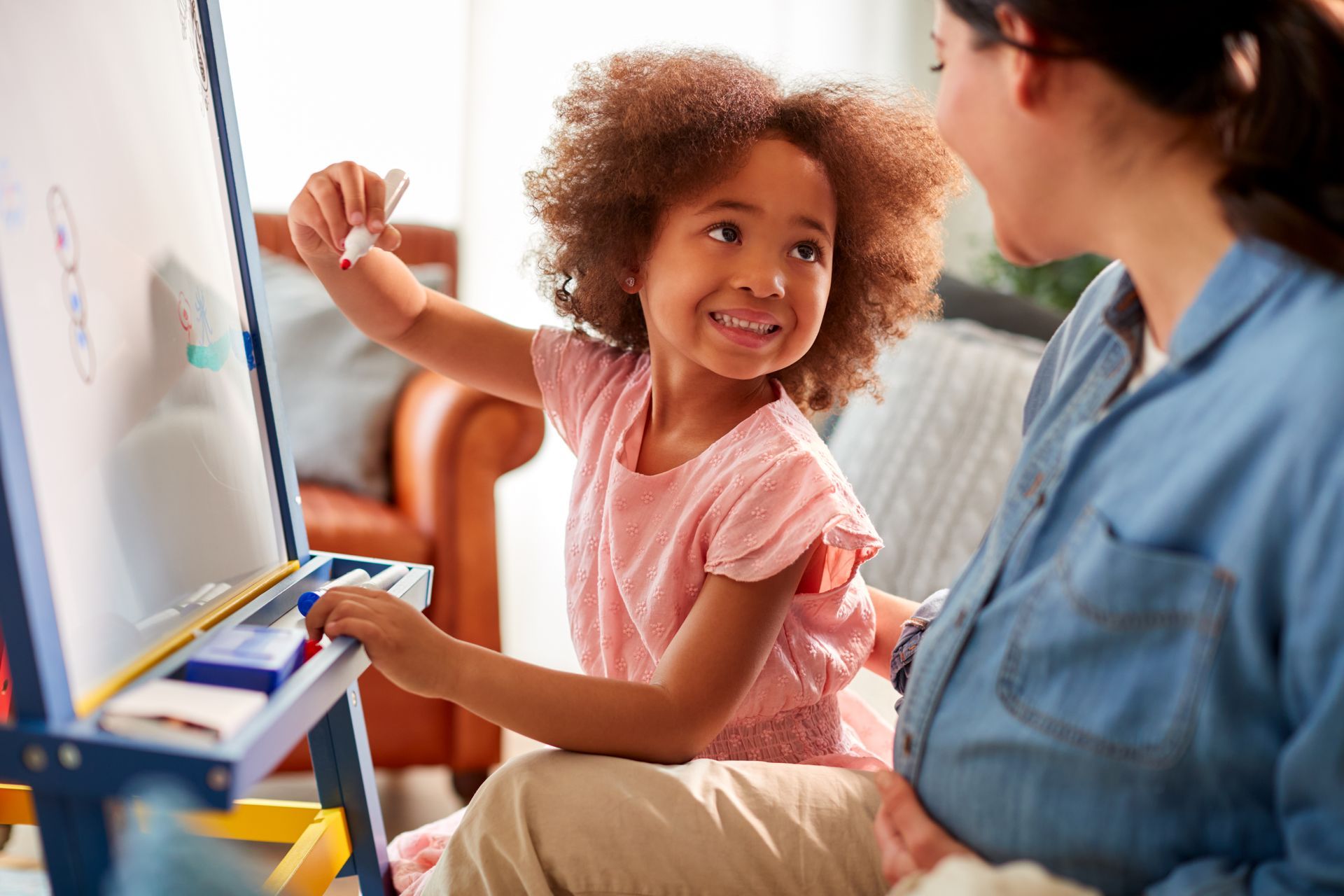 Girl drawing on an easel, smiling at a woman; indoor setting.