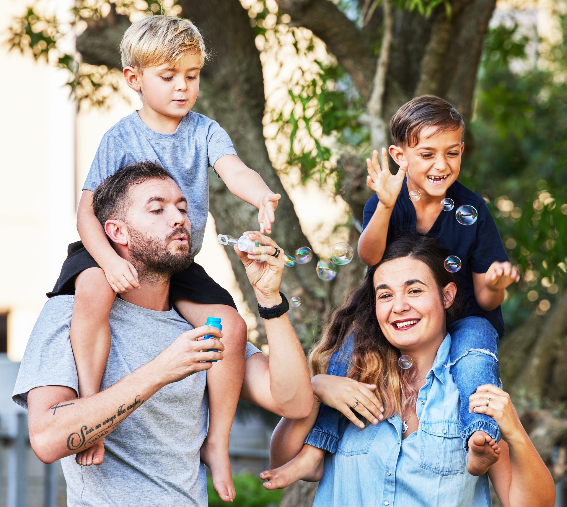 Family of four playing with bubbles outdoors; a child each on the parents' shoulders.