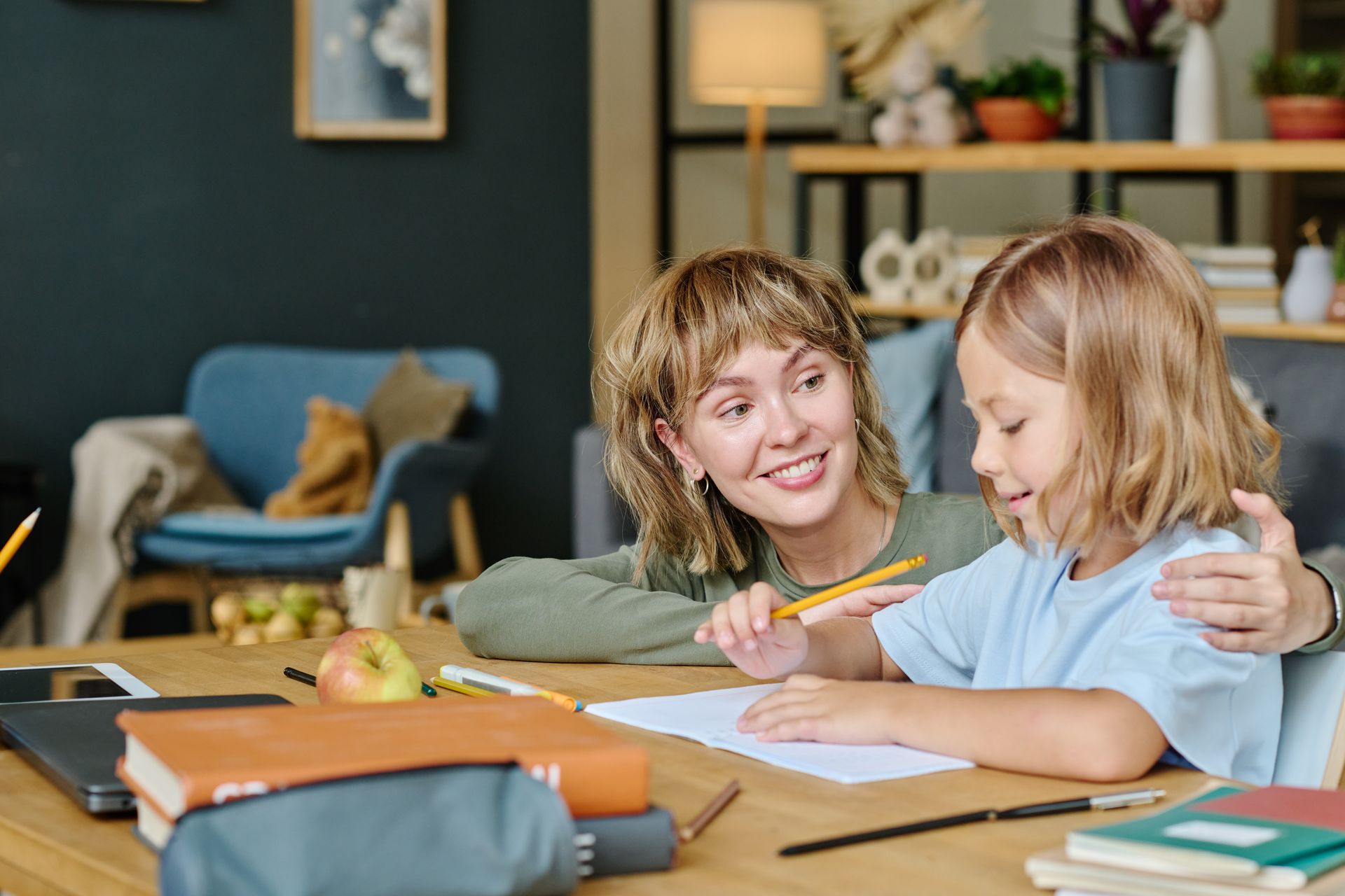 Woman helping child with homework at a table. Both are smiling, focused. Room with books, laptop, and fruit.