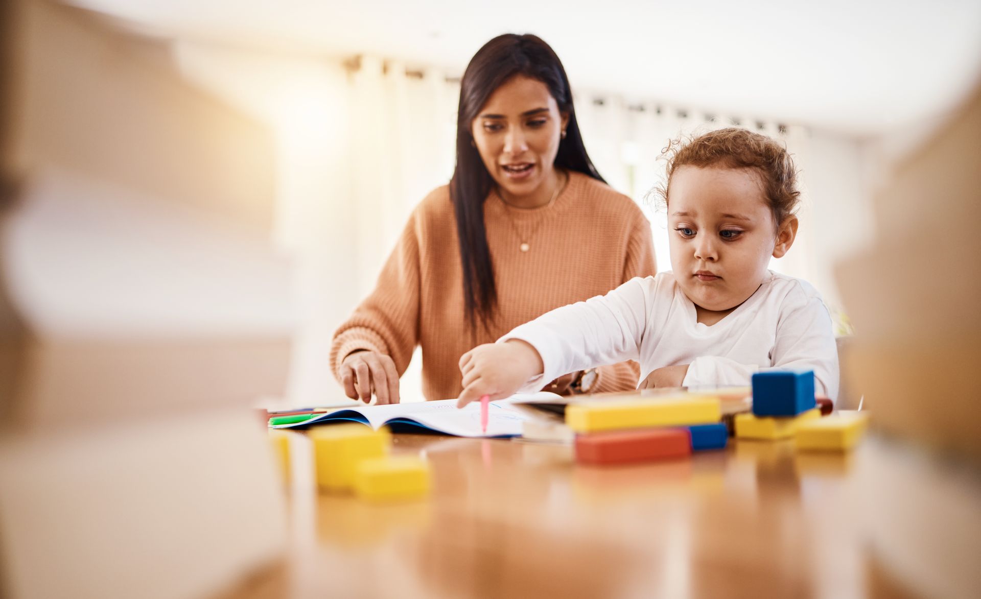 Woman assisting child with drawing at a table, blocks visible, natural light.