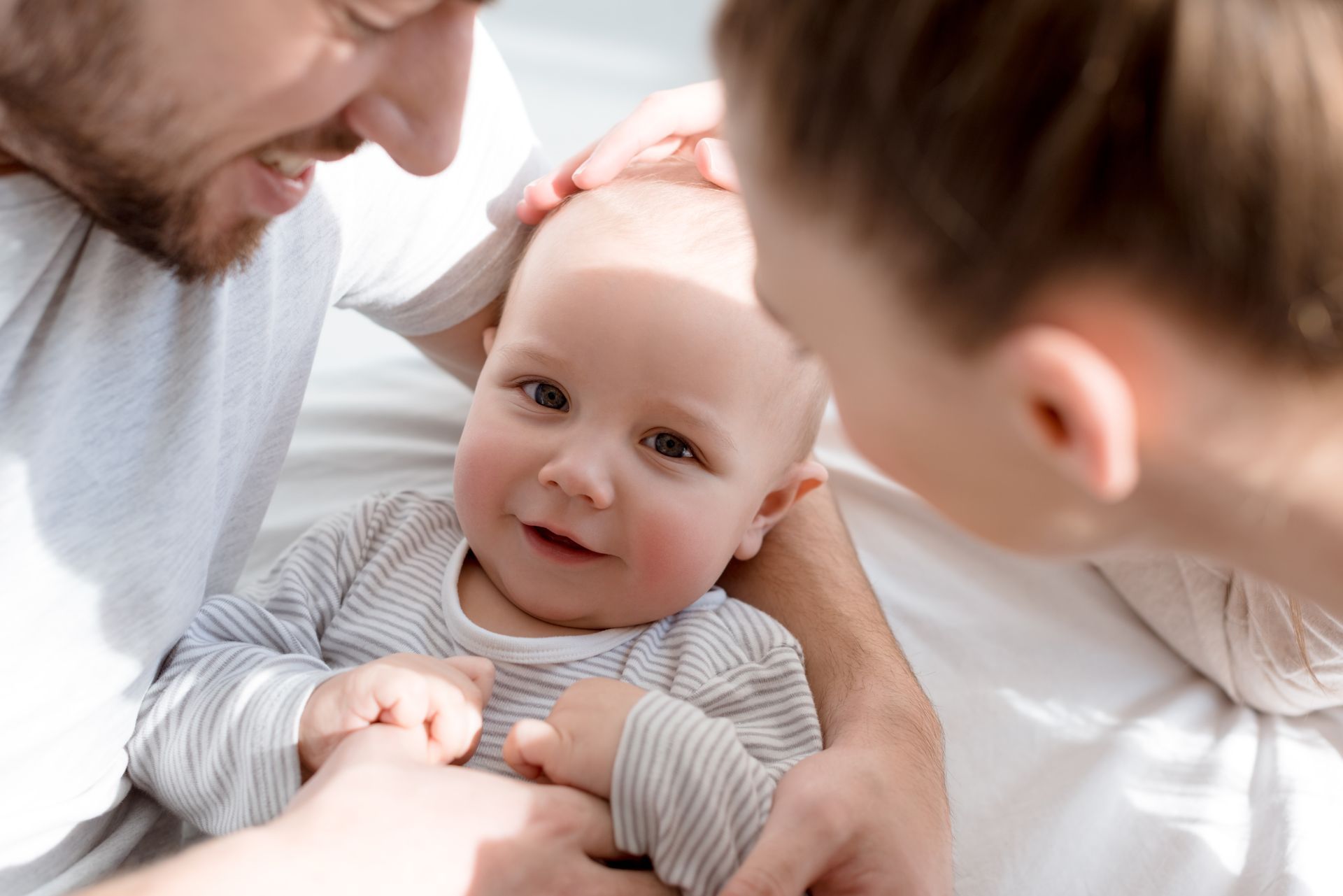 Parents smiling and touching a baby, who is lying down and looking at the camera.
