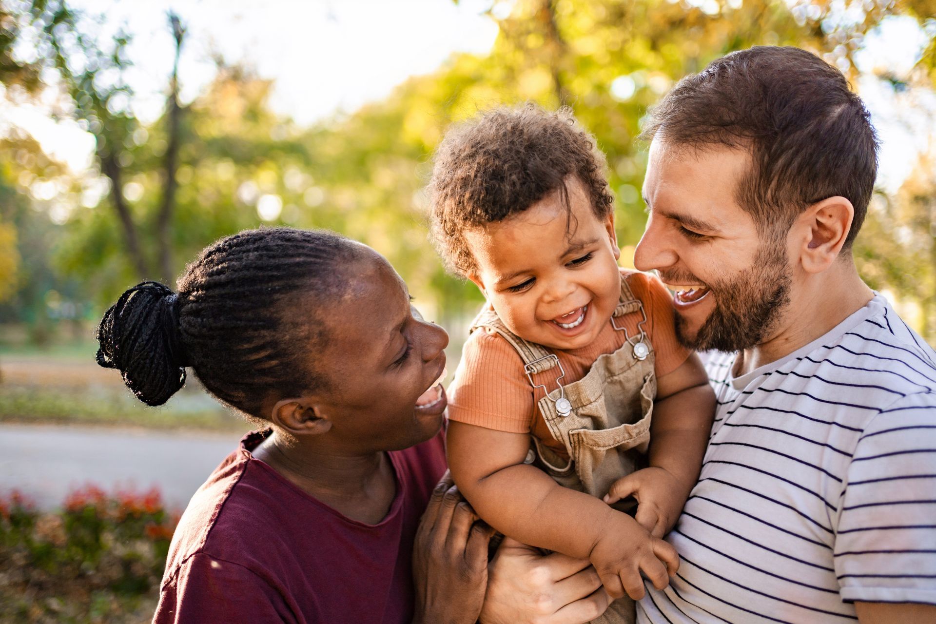 Parents smiling at and holding a child outdoors, trees in background.