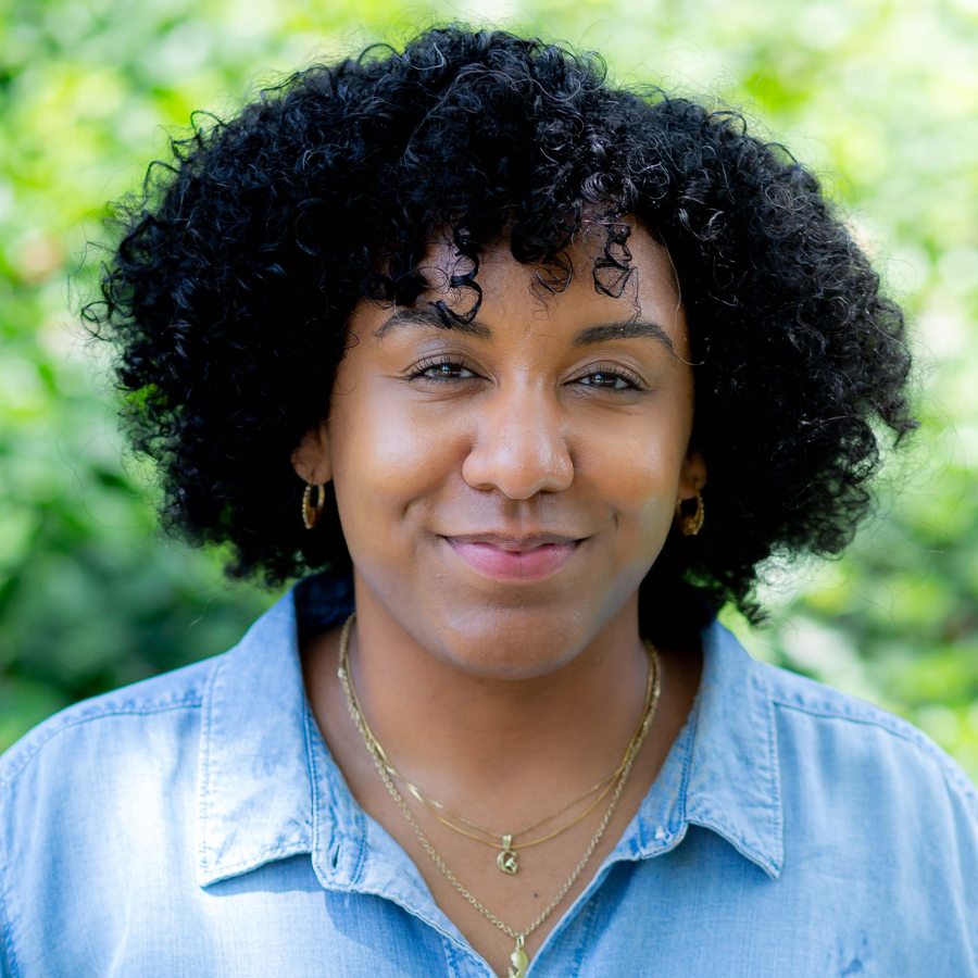 Woman with curly dark hair, smiling, wearing a cream-colored collared shirt, against a white wall.