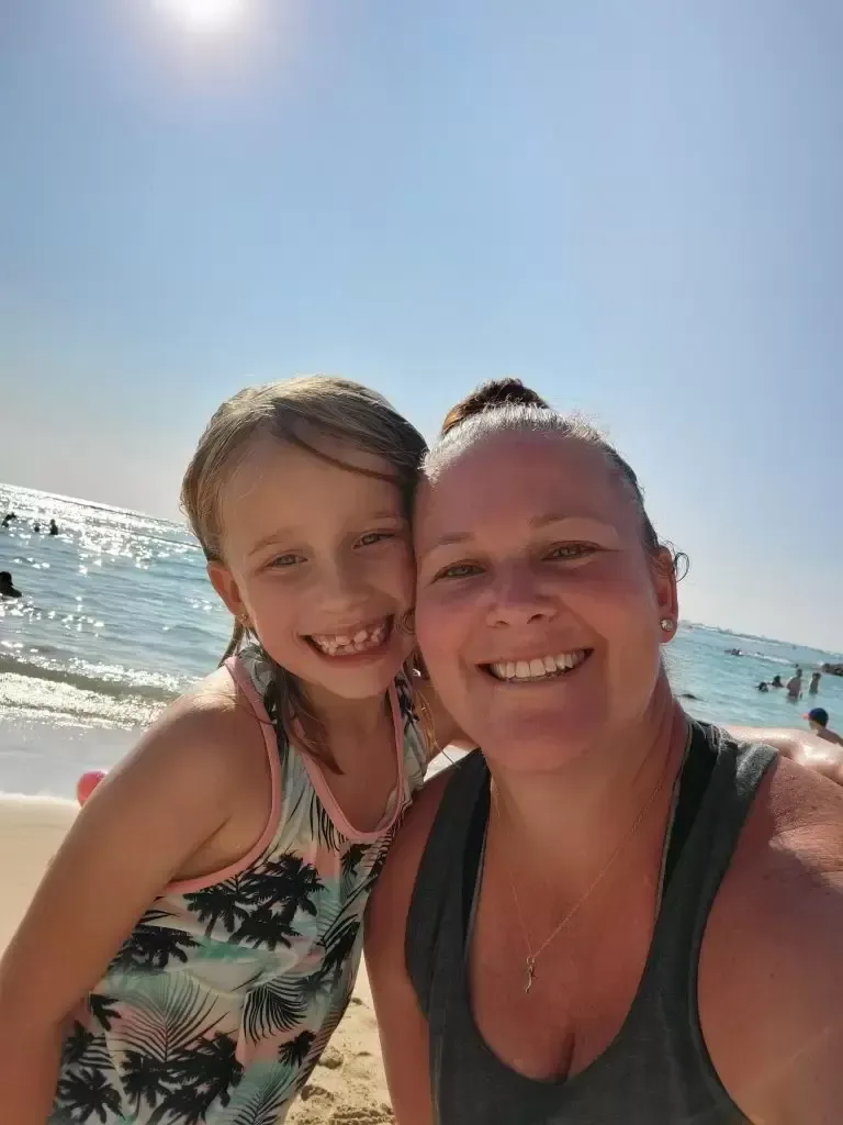 Woman and girl smile, taking selfie on beach; blue sky, ocean waves in background.