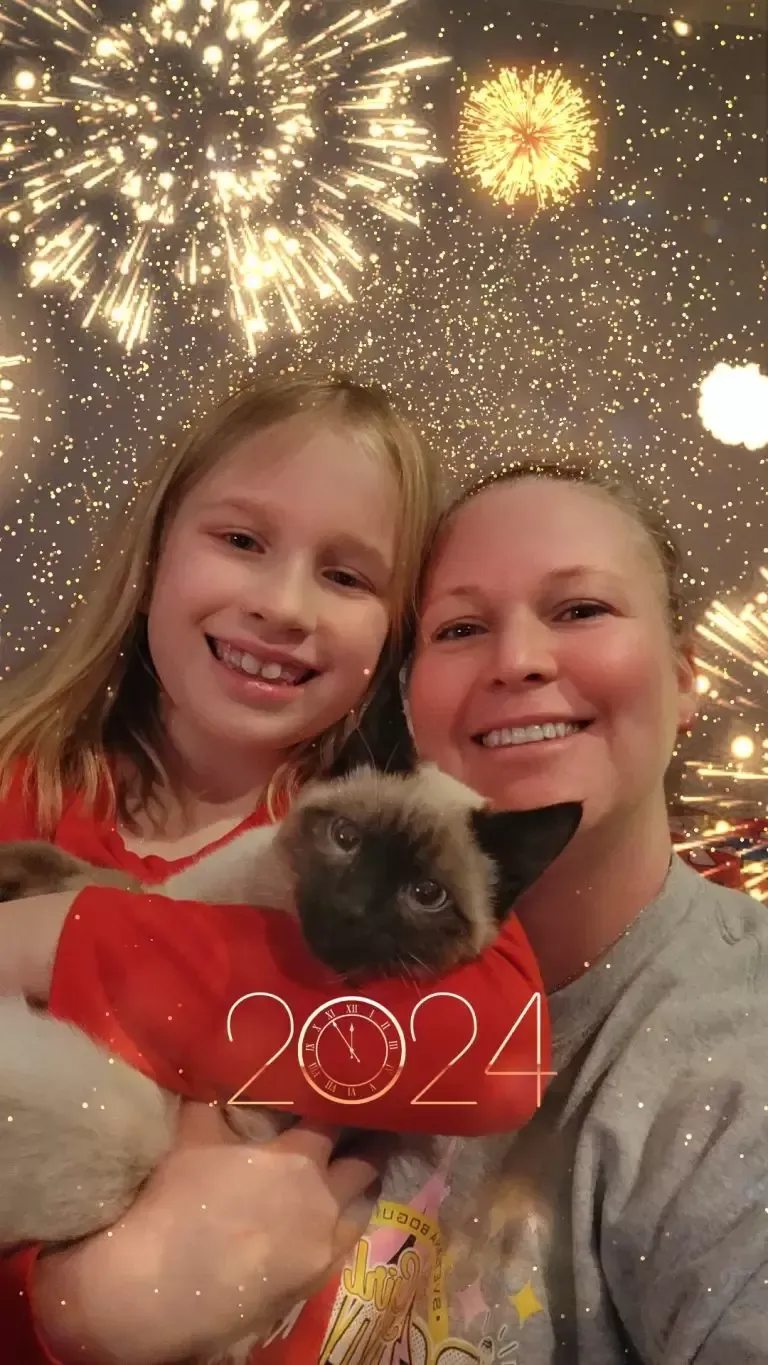 Woman and child smiling, holding a Siamese cat. Fireworks background with