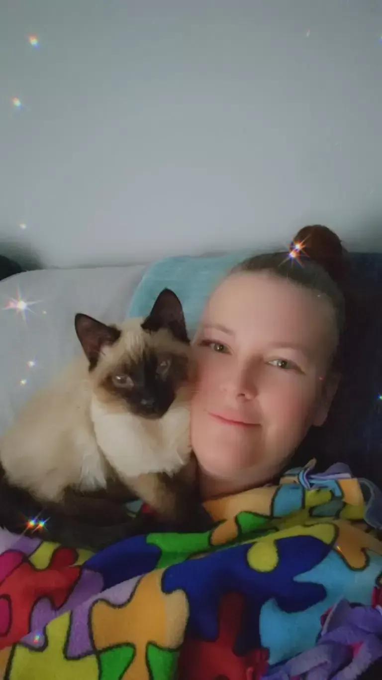 Woman with light skin and a cat with Siamese markings, snuggling under a colorful blanket.