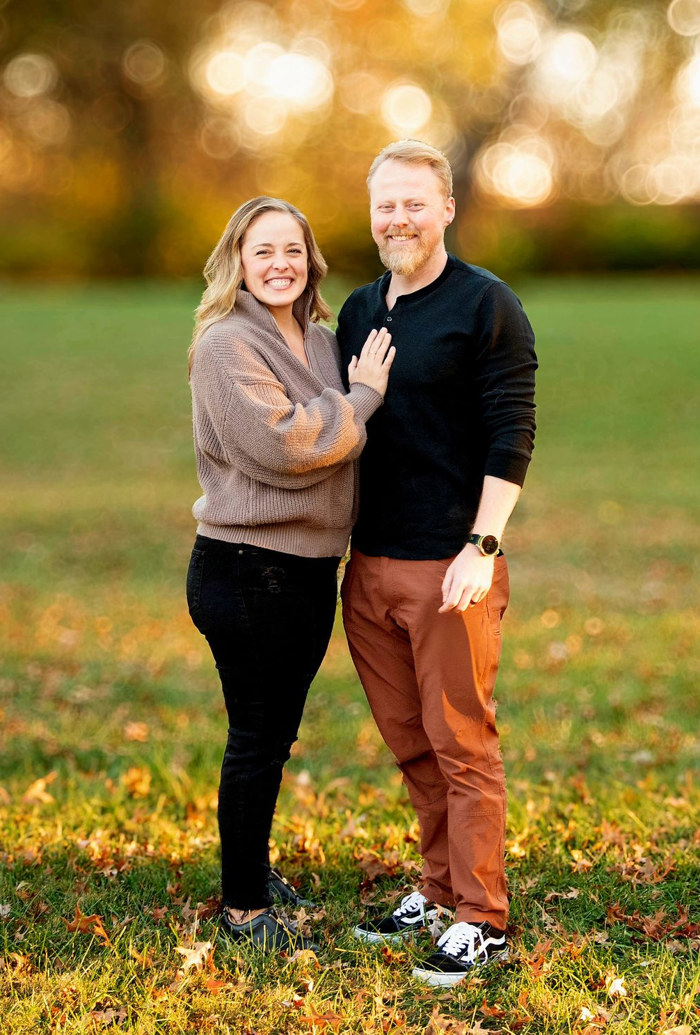 Couple smiling outdoors. Woman in blue dress, man in suit. Near a lamp post, tree, and foliage.