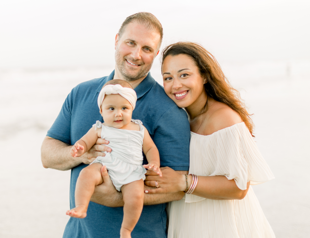 Family of three smiling at the beach; father holding baby, mother leaning in; sunset.