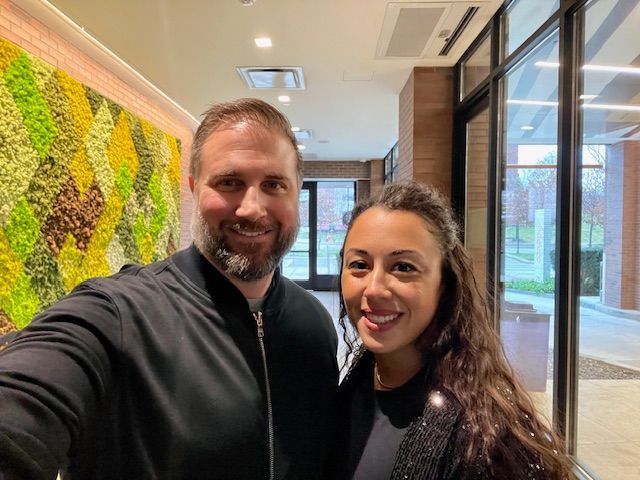 A man and woman smile, posing for a selfie indoors. A green and yellow wall and a glass door are visible.