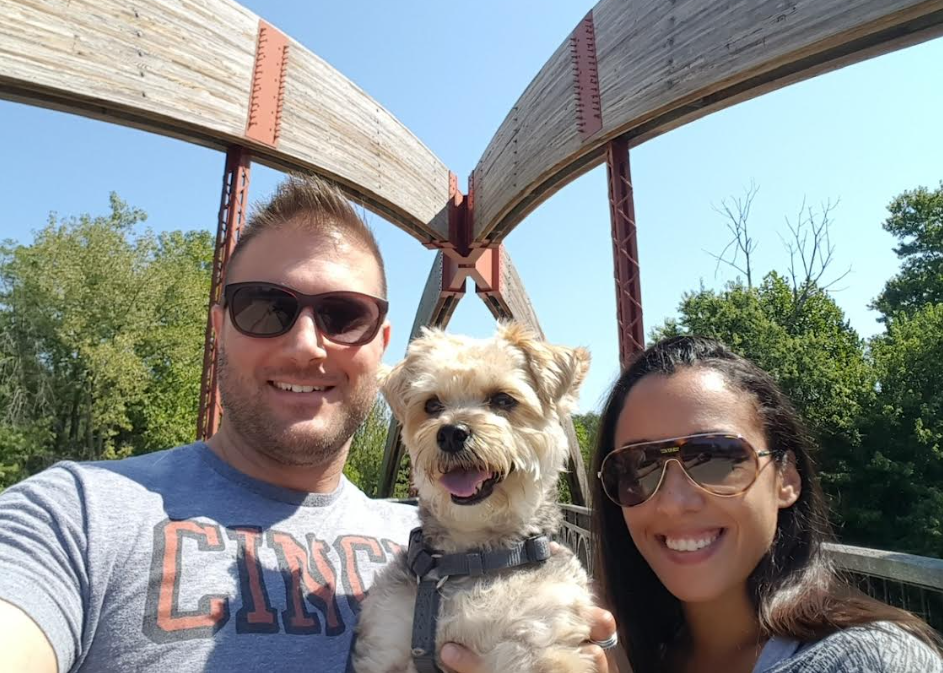 A couple and their dog smiling on a wooden bridge on a sunny day.