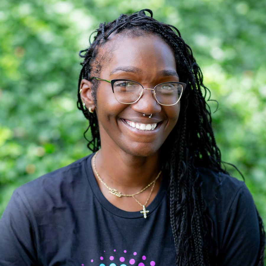 Smiling Black person with braids, glasses, nose ring, and gold necklaces against a blurred green backdrop.