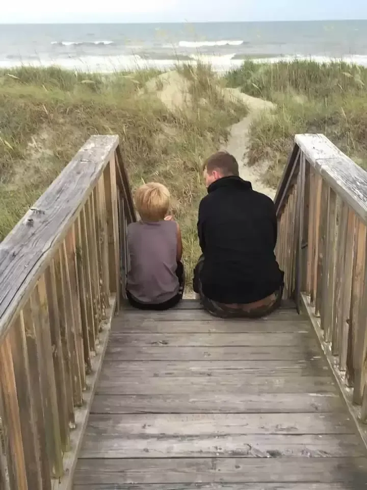 Man and child sitting on a wooden boardwalk, looking out at the ocean.