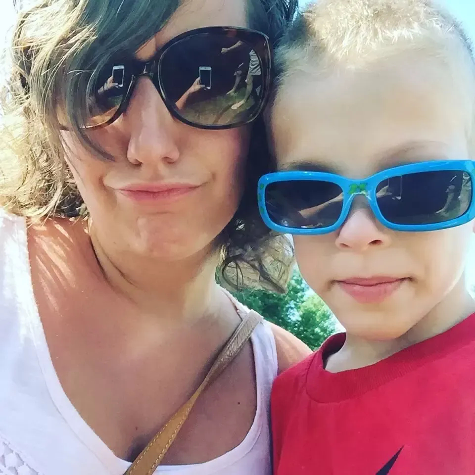 Woman and young boy wearing sunglasses pose for a selfie outdoors on a sunny day.