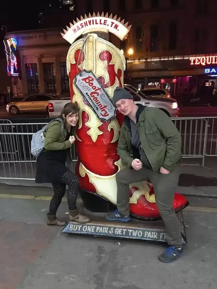 Couple poses with a giant boot sculpture in Nashville. Woman smiles, man flexes. Red, white, and black boot. Nighttime.