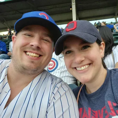 Man and woman smiling, wearing baseball caps, at a baseball game. Man in Cubs jersey, woman in a navy cap.
