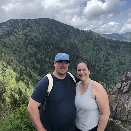 Man and woman pose together on a mountain overlooking green trees; man in blue hat, woman in white tank top.