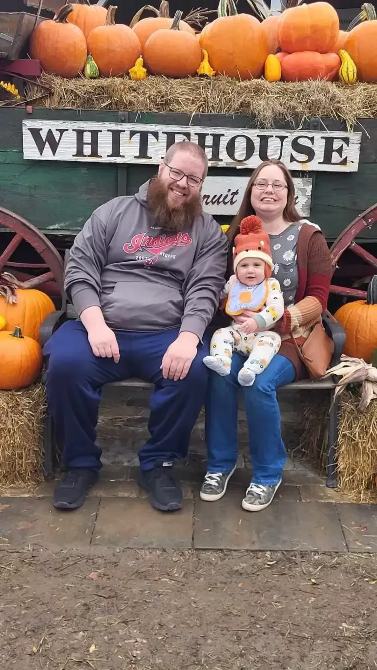 Family of three smiling in front of a White House sign with pumpkins. Baby held by a woman, man on the left.
