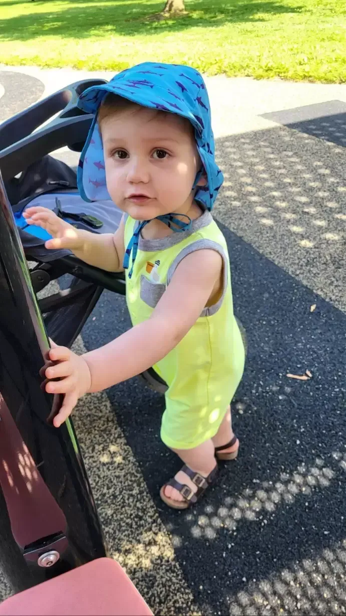 Baby in blue hat and yellow outfit standing at playground.