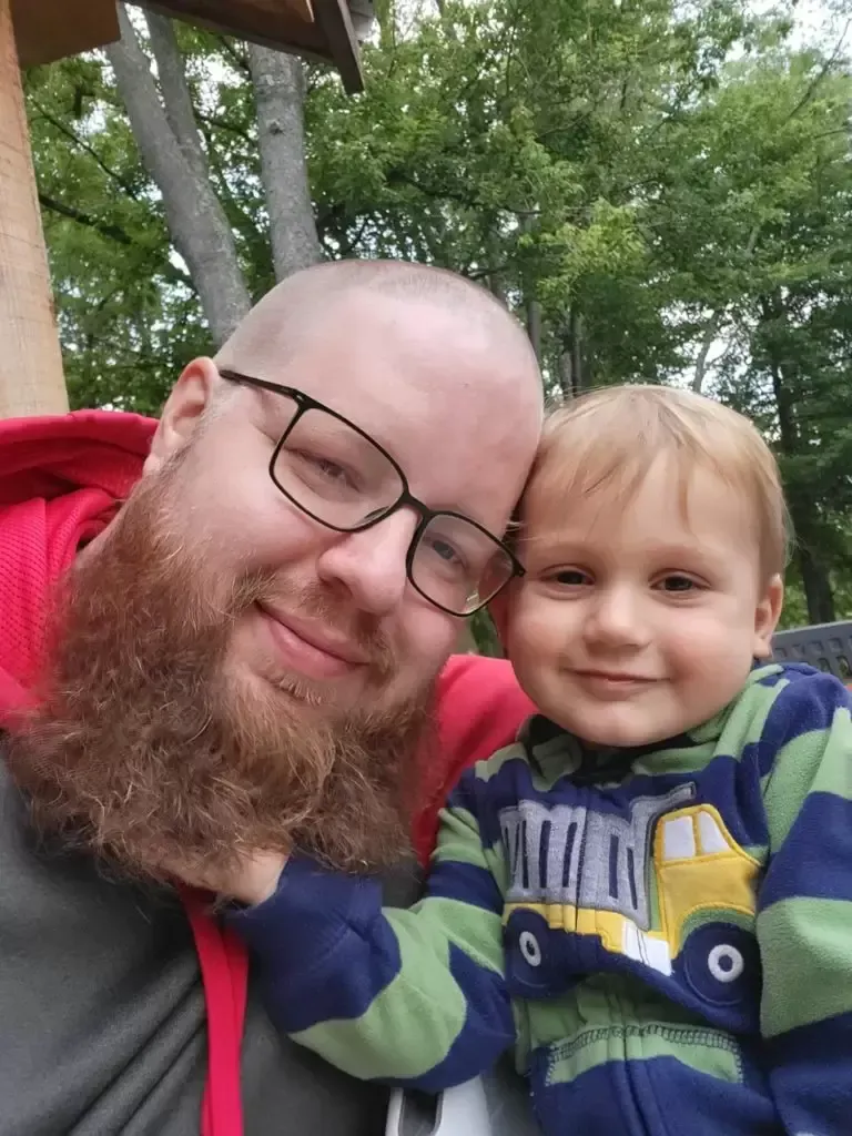 Man with a full beard and glasses smiles next to a smiling toddler in front of trees.
