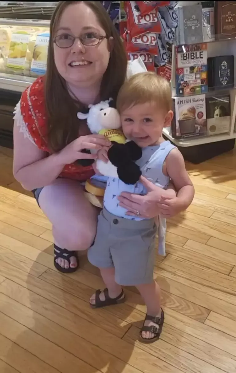 Woman and young child holding stuffed cow. Smiling woman kneels, child in blue shirt smiles, wooden floor.