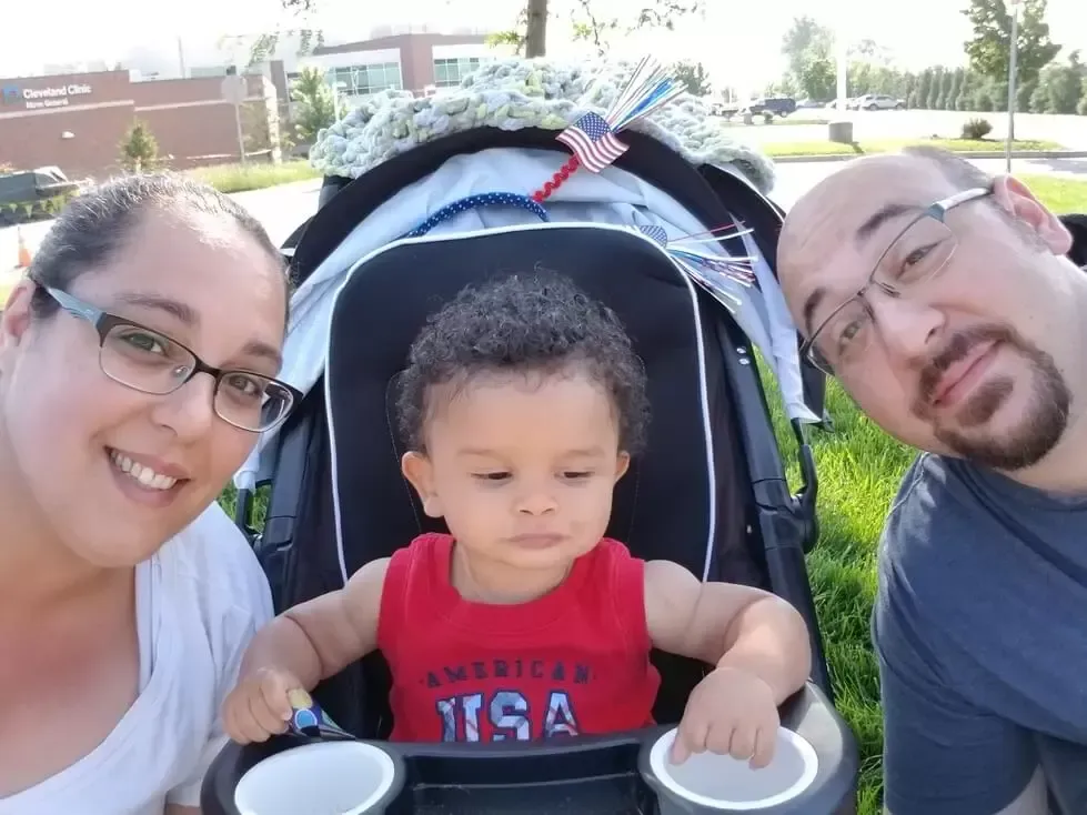 Family selfie outdoors: smiling parents flank baby in stroller; baby in red shirt looks down.