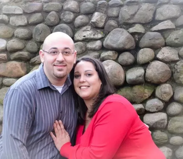 Couple smiling in front of a stone wall; man in blue shirt, woman in red sweater.