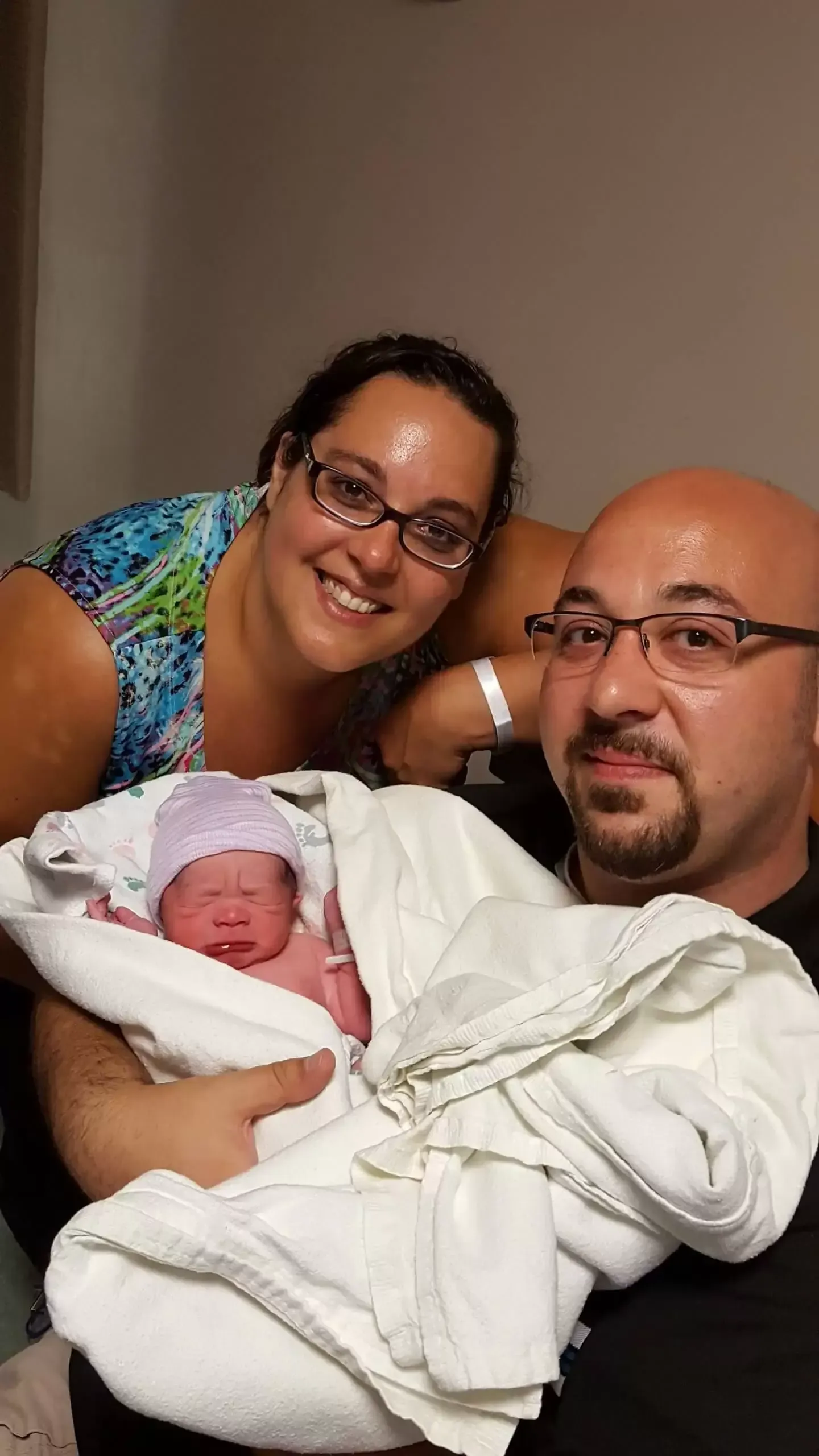 Parents holding newborn baby, hospital room setting, smiling, wrapped in blanket.