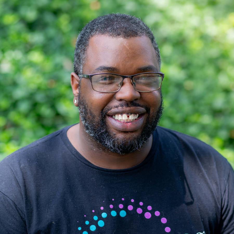 Man with glasses, dark skin, and beard smiles. Outdoors, blurred green background.