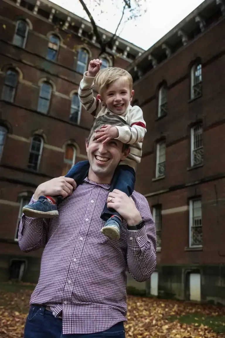 Man carrying a smiling child on his shoulders outdoors, in front of a brick building.