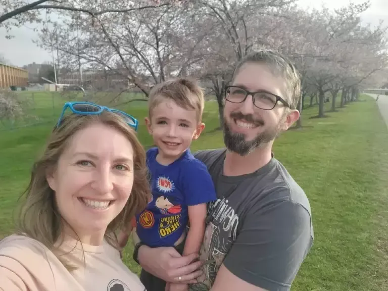 Family of three smiling, outdoors: woman, man holding child. Cherry trees in bloom in the background.