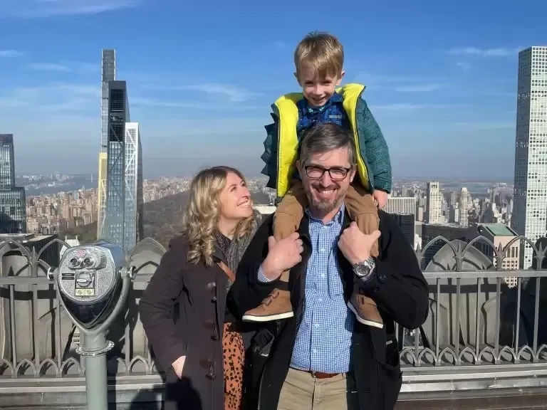 Family atop a skyscraper overlooking NYC. Father with son on shoulders, mother smiling, buildings in the distance.