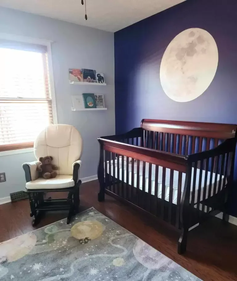 Nursery with dark wood crib, rocker, and moon mural on blue wall. Bookshelves and rug with space theme.