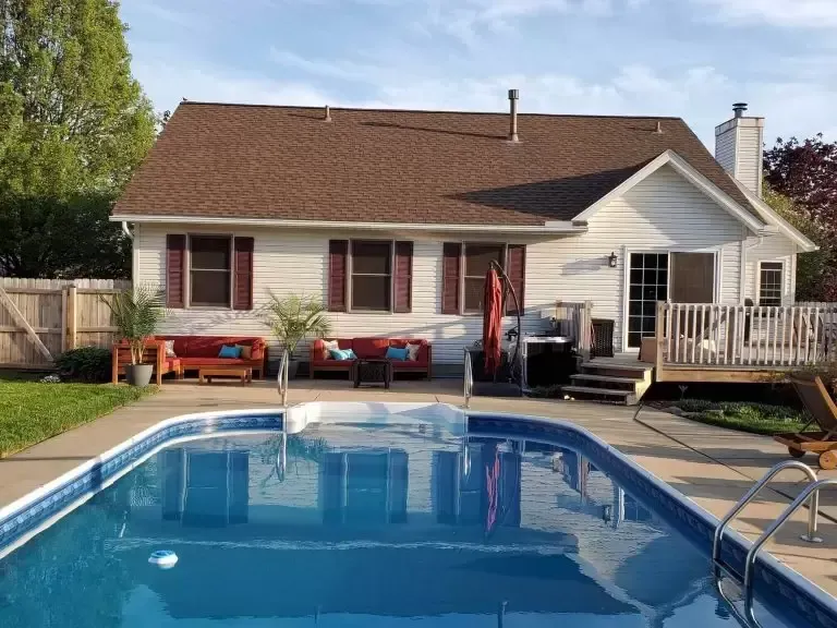 Backyard with pool, house with brown roof, red outdoor furniture, and a wooden deck.