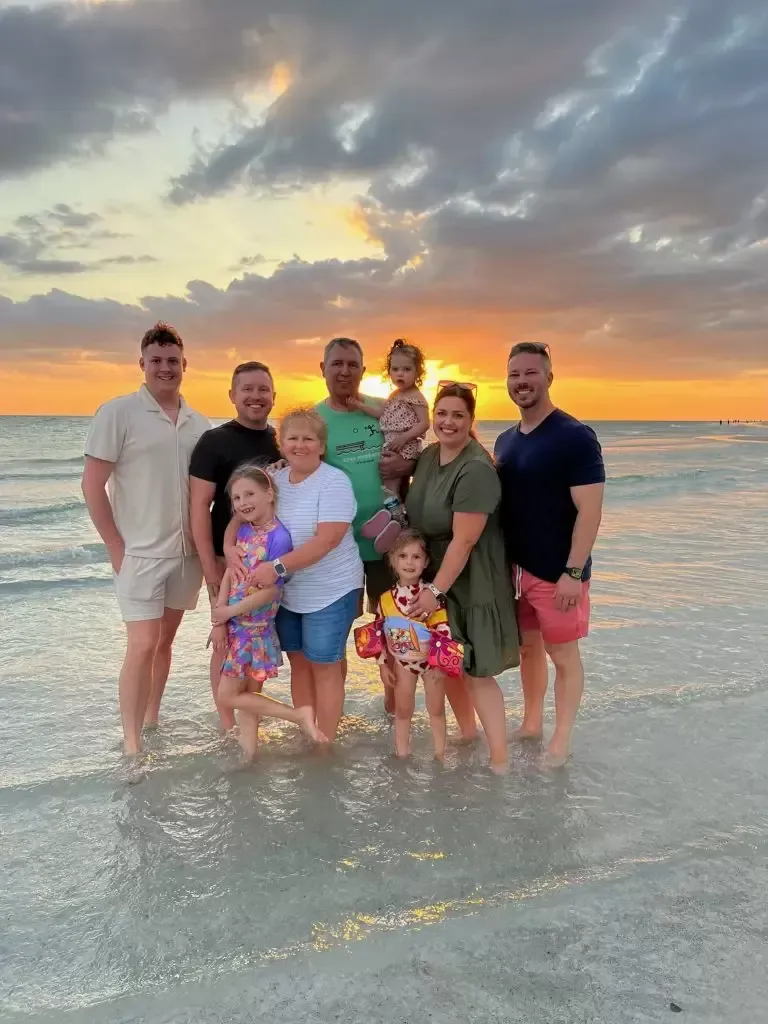 Family standing on a beach at sunset. People of varying ages in casual clothing.